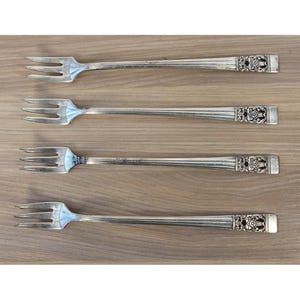 Pode incluir: Um conjunto de quatro garfos de cocktail em metal prateado, cada um com três dentes e um cabo longo e fino. As extremidades dos cabos são decoradas com um padrão floral em relevo, enquanto os cabos têm um design liso e ranhurado.