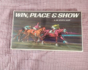 Könnte beinhalten: Vintage Brettspielbox "WIN, PLACE & SHOW" mit einer farbenfrohen Illustration eines Pferderennens mit Jockeys. Der Text "A 3M SPORTS GAME" ist zu sehen. Der Hintergrund ist ein rosa gesteppter Stoff.