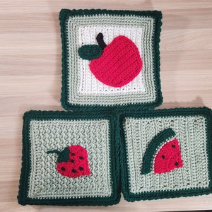 Puede incluir: Tres cuadrados de ganchillo con bordes verdes, cada uno con una fruta diferente: una manzana roja, una fresa roja y una sandía roja.