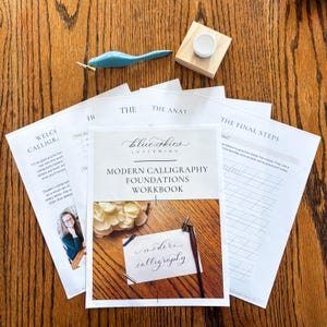 May include: A modern calligraphy workbook titled "Modern Calligraphy Foundations Workbook" with a blue pen holder, a wooden ink well, and practice sheets. The workbook cover features the text "Blue Skies Lettering". The image is on a wooden surface.