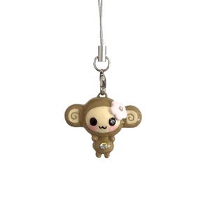 May include: A brown monkey charm attached to a silver-toned metal lobster clasp and a thin silver cord. The monkey has a cream-colored face, large black eyes, a small pink flower on its head, and a single clear rhinestone embedded on its belly.