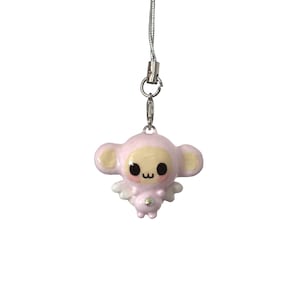 May include: A light pink charm featuring a stylized creature with large black eyes, a small mouth, and rounded ears. The figure has small white wings on its back and a single clear rhinestone on its chest, attached to a silver-colored cord.