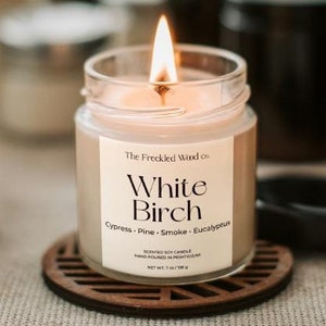 May include: A clear glass candle jar with a black label that reads "The Freckled Wood Co. White Birch Cypress - Pine - Smoke - Eucalyptus" and a lit candle inside. The candle is surrounded by other candle jars with black lids.