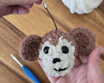 May include: A handmade crochet monkey face charm in brown and cream colors. The monkey has large black eyes, a black nose, and a smiling mouth. The charm is attached to a black string with a silver-colored clasp.