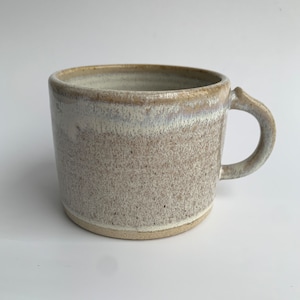 May include: A white ceramic mug with a speckled glaze and a light brown rim. The mug has a rounded handle.