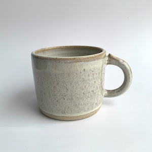 May include: A white ceramic mug with a speckled glaze and a brown rim. The mug has a rounded handle.