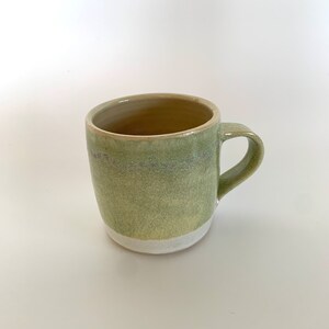 Könnte beinhalten: Eine grüne Keramiktasse mit weißem Rand. Die Tasse hat eine hellgrüne Glasur mit einem dunkleren grünen Band um die Mitte.