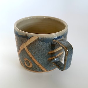 May include: A ceramic mug with a blue and brown glaze and a beige handle. The mug has a geometric pattern in beige.