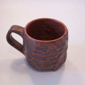 Könnte beinhalten: Eine kleine, braune Keramiktasse mit einem strukturierten, geometrischen Design. Die Tasse hat einen abgerundeten Griff und einen leicht ausgestellten Rand.