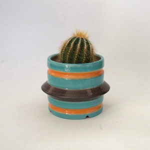 Puede incluir: Un pequeño cactus redondo en una maceta de cerámica turquesa con rayas naranjas y marrones. La maceta tiene un diseño único en capas, que añade un toque de estilo moderno. El cactus tiene espinas verdes y una parte superior amarilla.