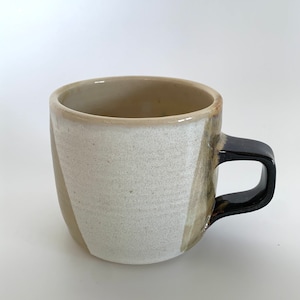 Könnte beinhalten: Eine weiße Keramiktasse mit einem schwarzen Henkel. Die Tasse hat einen braunen Überzug an der Seite und einen weißen Überzug an der Vorderseite.