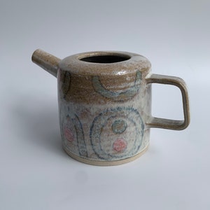 May include: A ceramic teapot with a light brown glaze and abstract geometric patterns in blue, green, and pink.