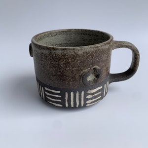 Könnte beinhalten: Eine braune Keramiktasse mit einem weißen geometrischen Muster am Boden. Die Tasse hat einen kleinen Henkel und eine leicht abgerundete Form.