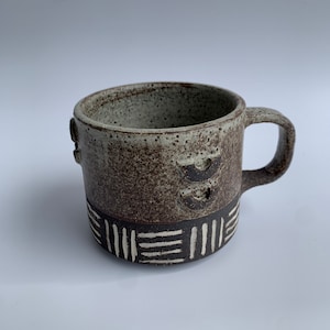 Könnte beinhalten: Eine braune und weiße Keramiktasse mit geometrischem Muster. Die Tasse hat eine gesprenkelte Glasur und einen Henkel.