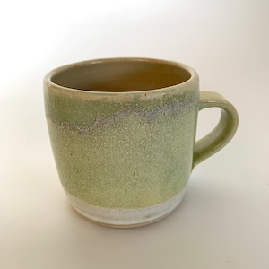 Könnte beinhalten: Eine grüne Keramiktasse mit weißem Rand und einem weißen Band am Boden. Die Tasse hat eine gesprenkelte Glasur.