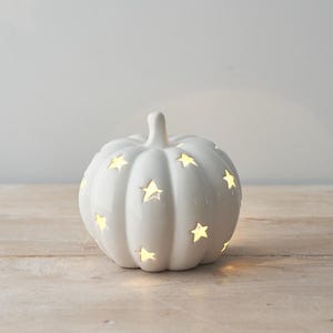 Può includere: Una lanterna in ceramica bianca a forma di zucca con finitura lucida, caratterizzata da molteplici intagli a forma di stella che lasciano filtrare una luce calda e soffusa dall'interno. L'oggetto presenta un corpo arrotondato e costolato.