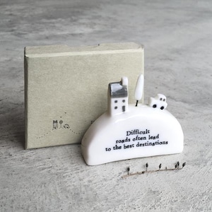 May include: A white ceramic decorative piece with a house, tree, and car design, and the text "Difficult roads often lead to the best destinations." A small, light brown box is behind the ceramic piece. A small metal hook rack is in front of the ceramic piece.
