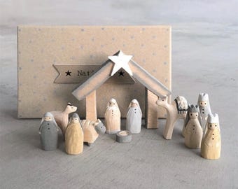 May include: A wooden nativity scene set with a star-topped stable, figures of the Holy Family, animals, and the Three Wise Men. The figures are painted in muted tones of gray, beige, and white. The backdrop is a box with a star design.
