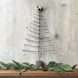 Op de afbeelding: Een rustieke kerstboom gemaakt van donker draad, bekroond met een houten ster. De boom staat op een houten voet tegen een lichte houten achtergrond. Groene bladeren en kleine witte bloemen sieren de basis.