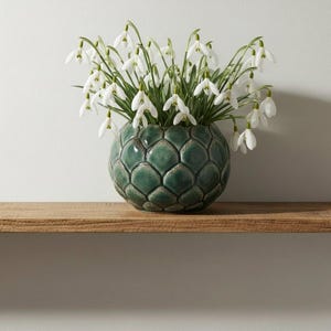 Puede incluir: Un jarrón de cerámica verde azulado con un patrón escamoso contiene un ramo de campanillas de invierno blancas. El jarrón está sobre una estantería de madera, creando una presentación sencilla. Las flores tienen pétalos blancos y tallos verdes. El jarrón mide unos 20 cm de diámetro.