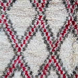 May include: White, gray, and red Moroccan rug with a geometric pattern. The rug is made of wool and has a soft, fluffy texture.