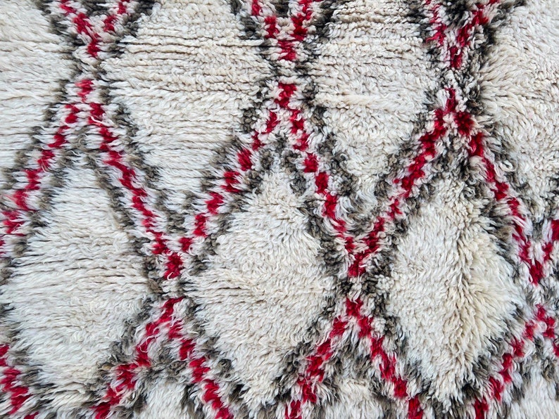 May include: White, gray, and red Moroccan rug with a geometric pattern. The rug is made of wool and has a soft, fluffy texture.