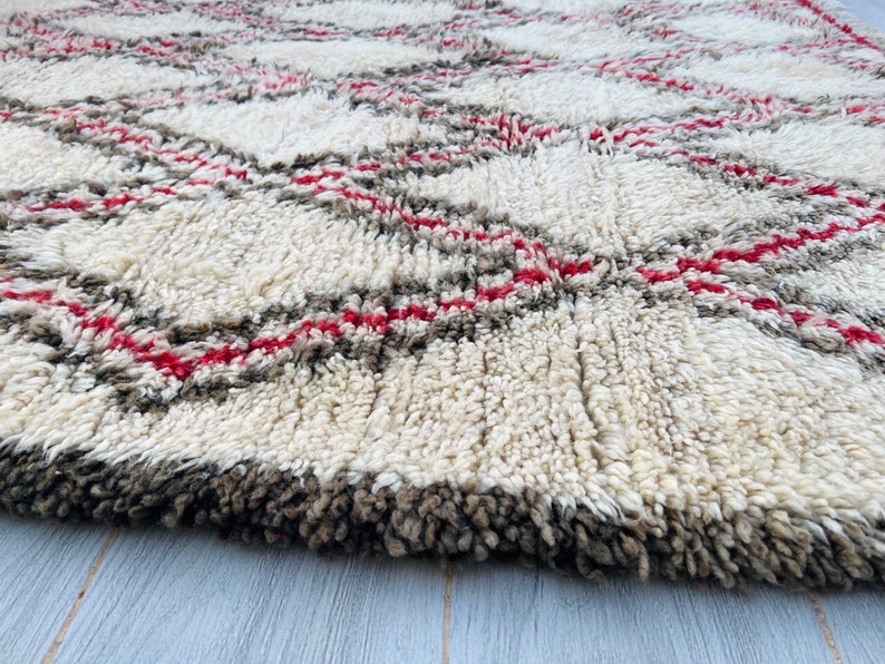 May include: A white, fluffy rug with a geometric pattern of red and gray lines. The rug has a thick pile and is made of wool.