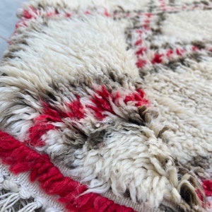 May include: Close-up of a white and gray Moroccan rug with red accents. The rug has a fringe border and a soft, fluffy texture.