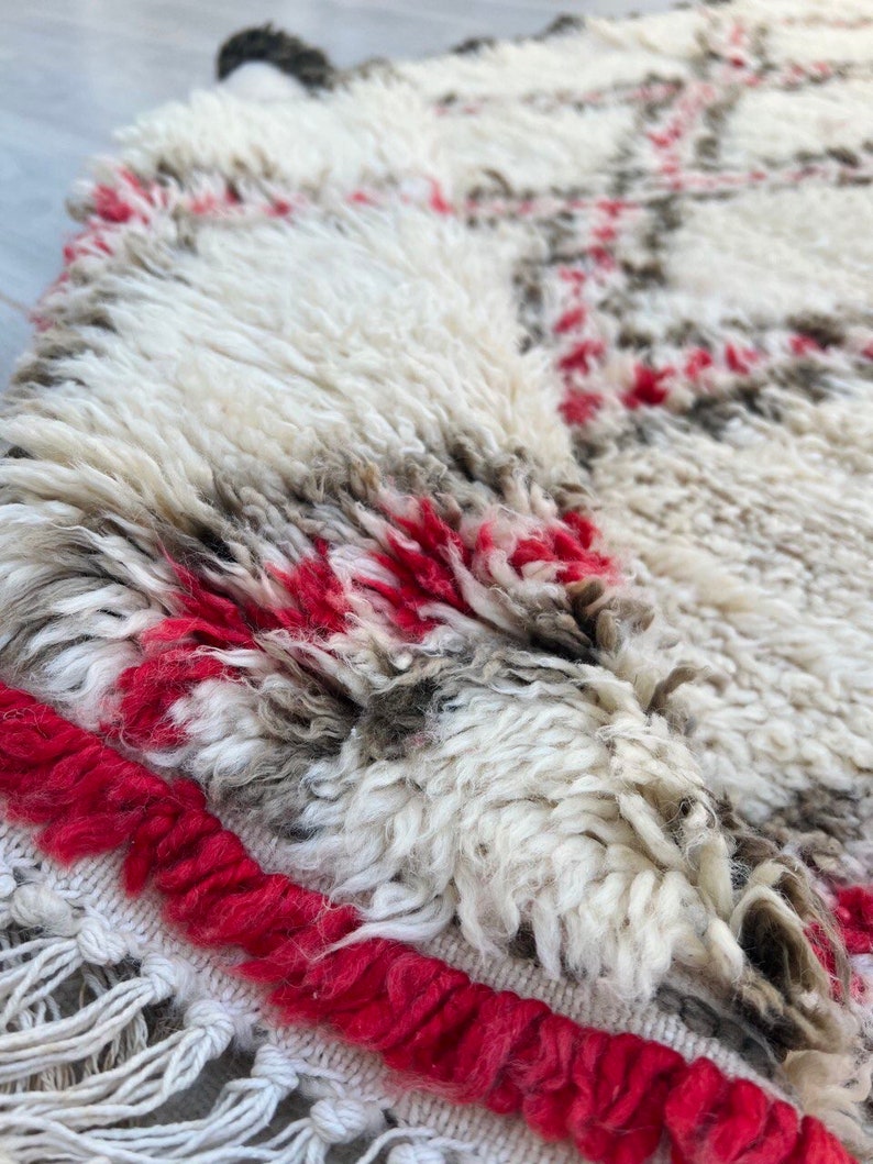 May include: Close-up of a white and gray Moroccan rug with red accents. The rug has a fringe border and a soft, fluffy texture.