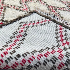 May include: Close-up of a white, gray, and red patterned rug with a fringe edge. The rug has a geometric design with stripes and diamonds.