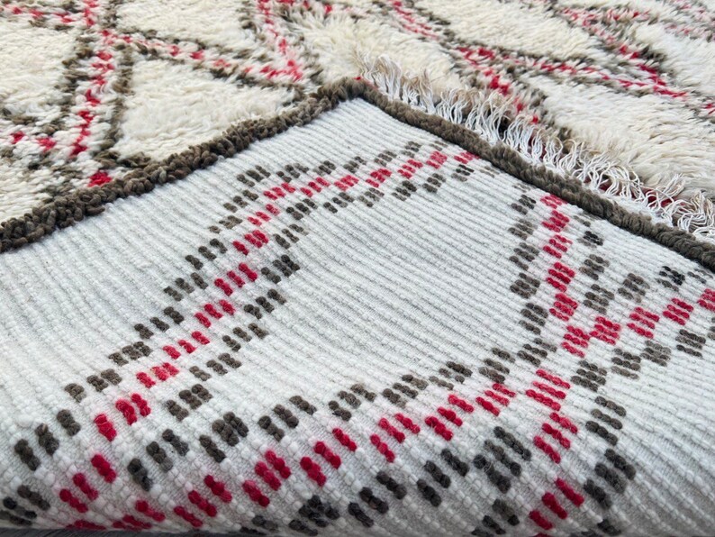 May include: Close-up of a white, gray, and red patterned rug with a fringe edge. The rug has a geometric design with stripes and diamonds.