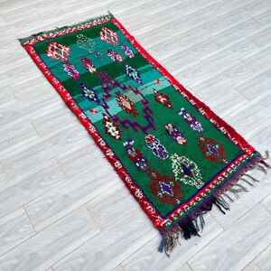Puede incluir: Una alfombra marroquí verde y roja con un patrón geométrico tradicional. La alfombra tiene un borde de flecos y está hecha de lana.