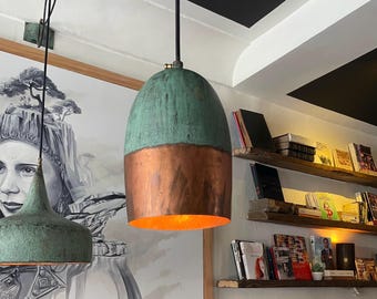 May include: A copper and verdigris pendant light fixture hangs from the ceiling. The upper portion is a weathered green, while the lower section is a warm copper color. The light fixture is in a room with bookshelves and a mural.