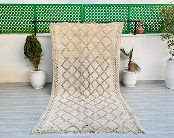 May include: A white Moroccan rug with a geometric pattern in black and brown. The rug is hanging on a clothesline against a green lattice background.