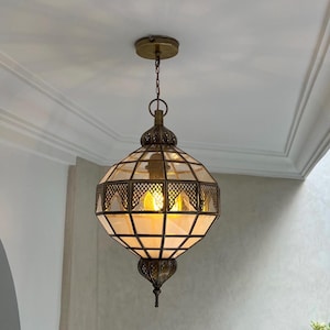 May include: An ornate pendant light fixture in an antique style. The brass-coloured metal frame has a geometric design with clear and amber-coloured glass panels. The light hangs from a chain and is suspended from a brass-coloured ceiling mount.