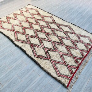 May include: A white Moroccan rug with a diamond pattern in red and black. The rug has a fringe border.