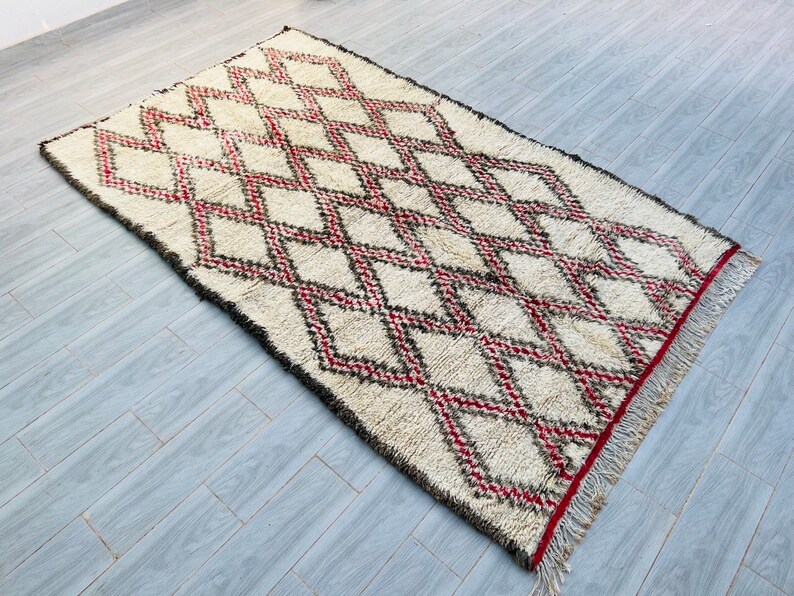 May include: A white Moroccan rug with a diamond pattern in red and black. The rug has a fringe border.
