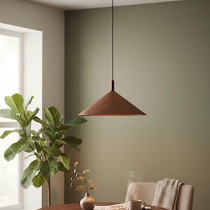 May include: A contemporary pendant light with a conical shade in a copper finish, suspended from a black cable. The light hangs above a dark wooden table. The room has a green wall and a large indoor plant, creating a warm atmosphere.