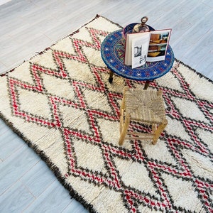 May include: A white, fluffy rug with a diamond pattern in red and black. The rug is on a tiled floor and there is a small wooden stool and a blue and white table with a book on top.
