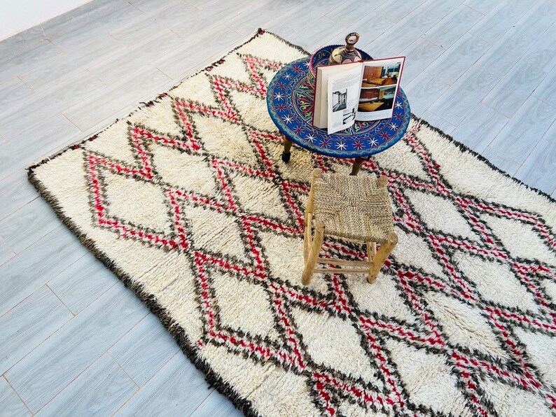 May include: A white, fluffy rug with a diamond pattern in red and black. The rug is on a tiled floor and there is a small wooden stool and a blue and white table with a book on top.
