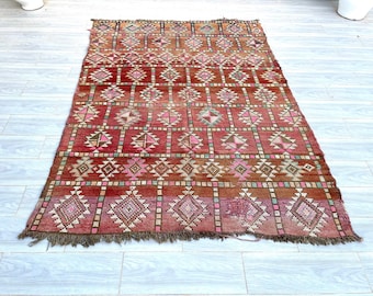 May include: A vintage Moroccan rug with a geometric pattern in shades of red, pink, and brown. The rug has a fringed edge and is woven with a variety of textures.