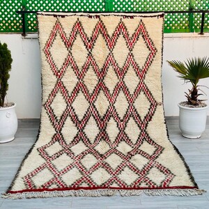 May include: A white, red, and black Moroccan rug with a diamond pattern. The rug has a fringe border and is laid out on a tiled floor.