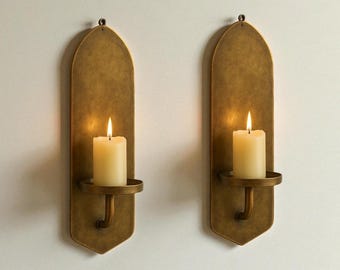 May include: Two wall-mounted candle holders with a brass finish. Each holder features a long, arched backplate, a circular candle cup, and a lit, ivory-colored pillar candle. The candle flames cast a warm glow.