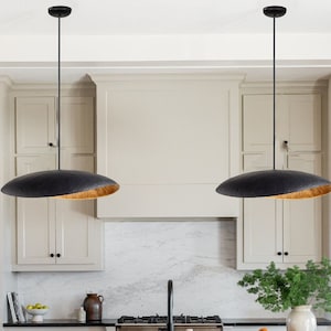 May include: Two black pendant lights with a textured surface and a gold interior hang from the ceiling. The lights are suspended by thin black cords. The kitchen features white cabinets and a marble countertop.