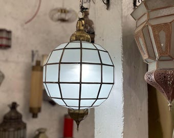 May include: A white and brass geometric pendant light with a globe shape. The light has a grid pattern and is hanging from a chain. Other decorative lights are in the background.