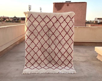 May include: A white and red Moroccan rug with a diamond pattern. The rug is hanging on a wall and has a fringe border.