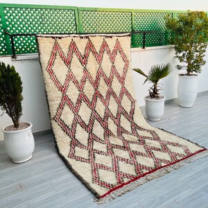 May include: A white and red Moroccan rug with a diamond pattern. The rug is hanging on a wall and has a fringe border.