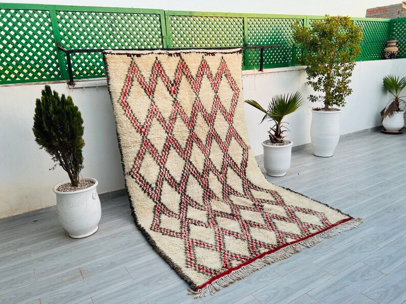 May include: A white and red Moroccan rug with a diamond pattern. The rug is hanging on a wall and has a fringe border.