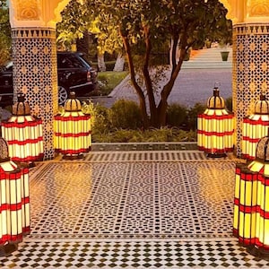 May include: Ornate, illuminated lanterns with red and yellow stained glass panels are arranged on a patterned tile floor. The lanterns are in a courtyard setting, with decorative columns and a glimpse of a car in the background.