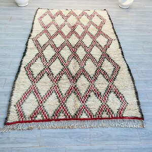 May include: A white and red Moroccan rug with a diamond pattern. The rug has a fringe border.
